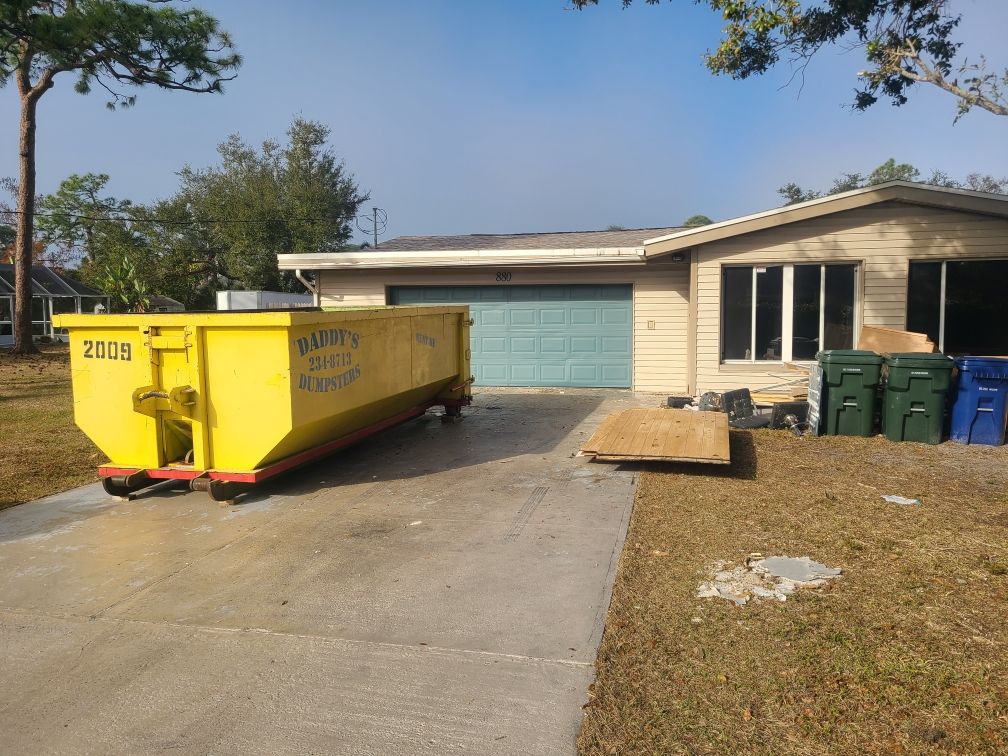 20-yard yellow dumpster in a residential driveway beside a house and garage, with trash bins nearby.