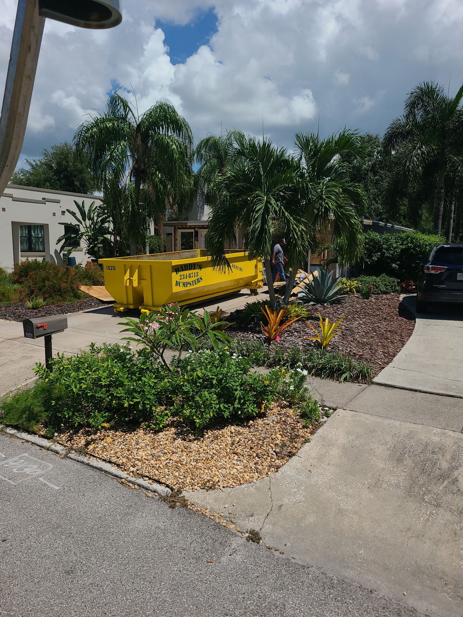 Yellow dumpster beside landscaped planting beds and a sidewalk on a sunny day