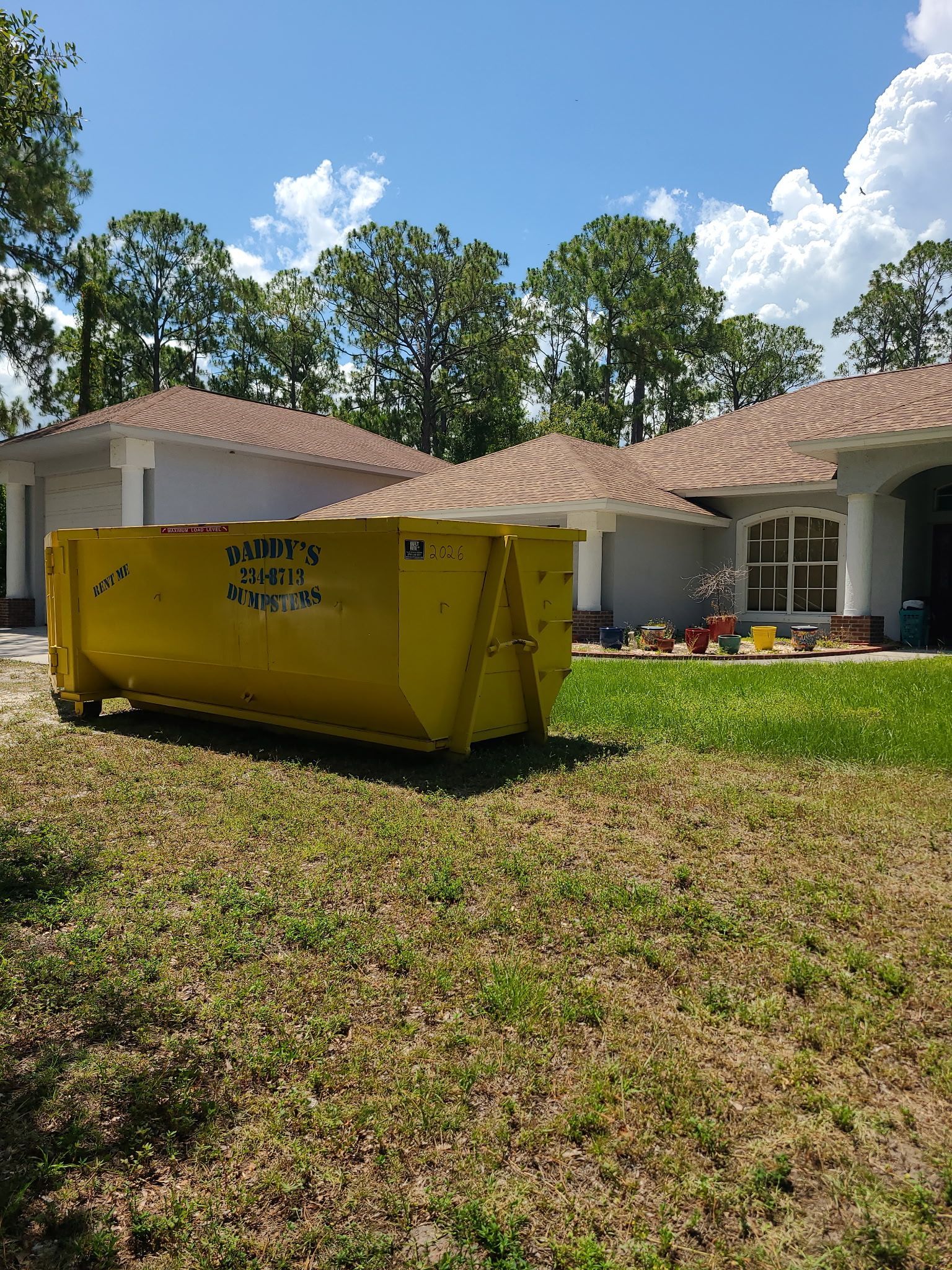20-yard yellow dumpster in a grassy yard in front of a white house with red roof tiles under a blue sky.