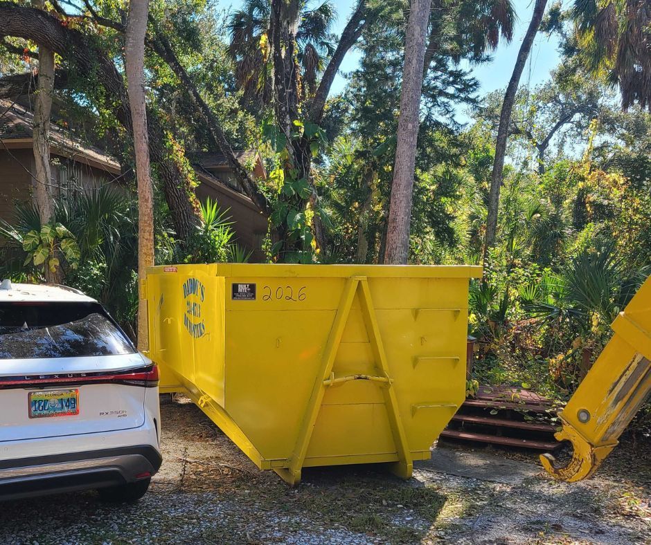 20-yard bright yellow dumpster beside a white SUV in a wooded yard, with construction equipment at right.