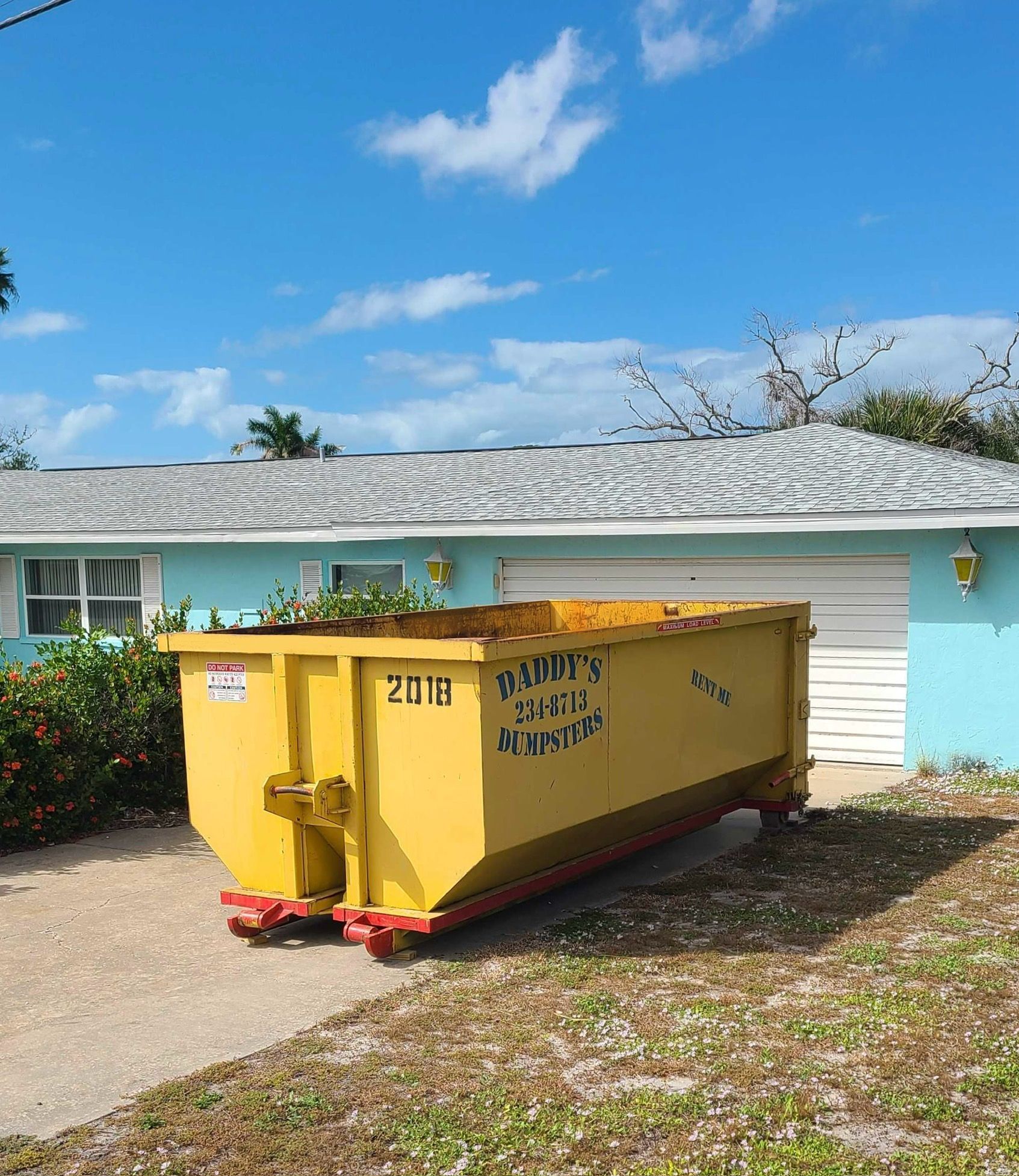 20-yard yellow dumpster in a driveway beside a light blue house under a clear sky.