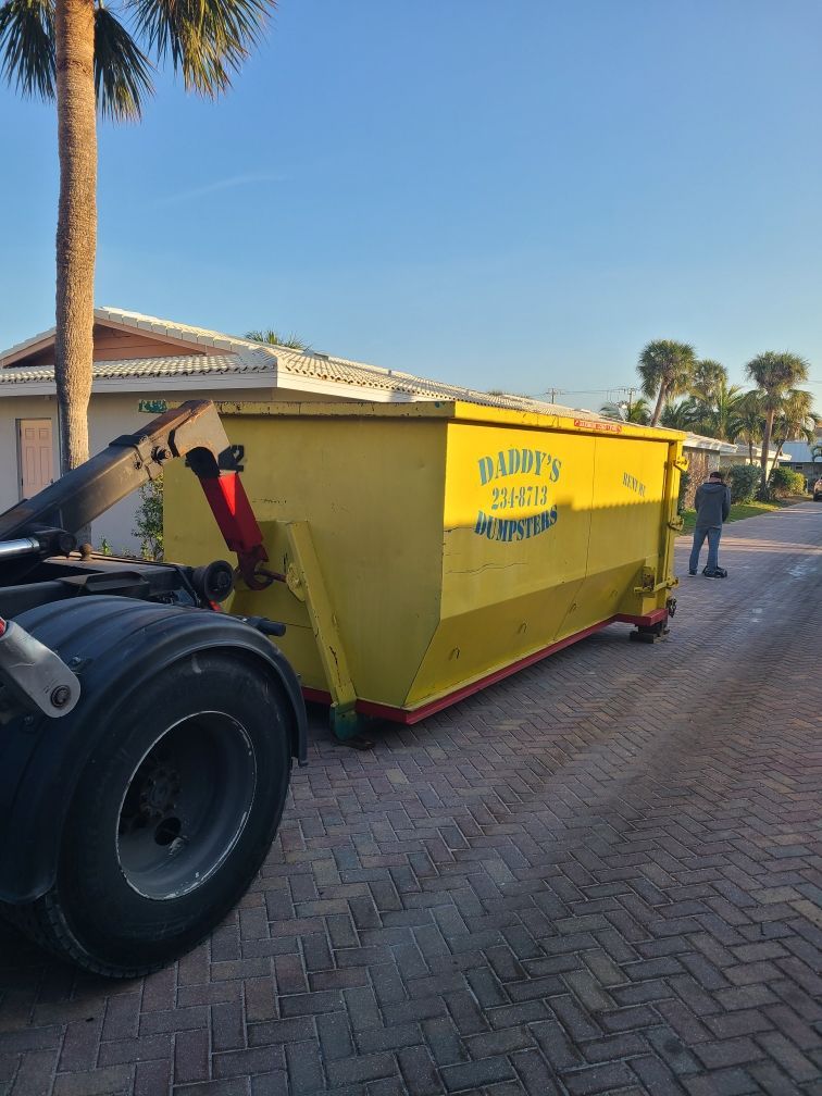 Yellow dumpster trailer on a brick driveway, with a worker standing nearby under a clear blue sky.