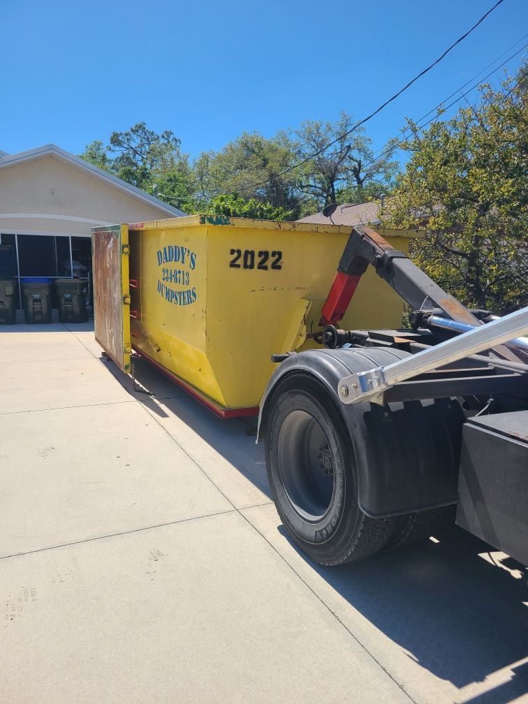 20-yard yellow construction dumpster beside a garage on a sunny driveway, hitched to a truck.