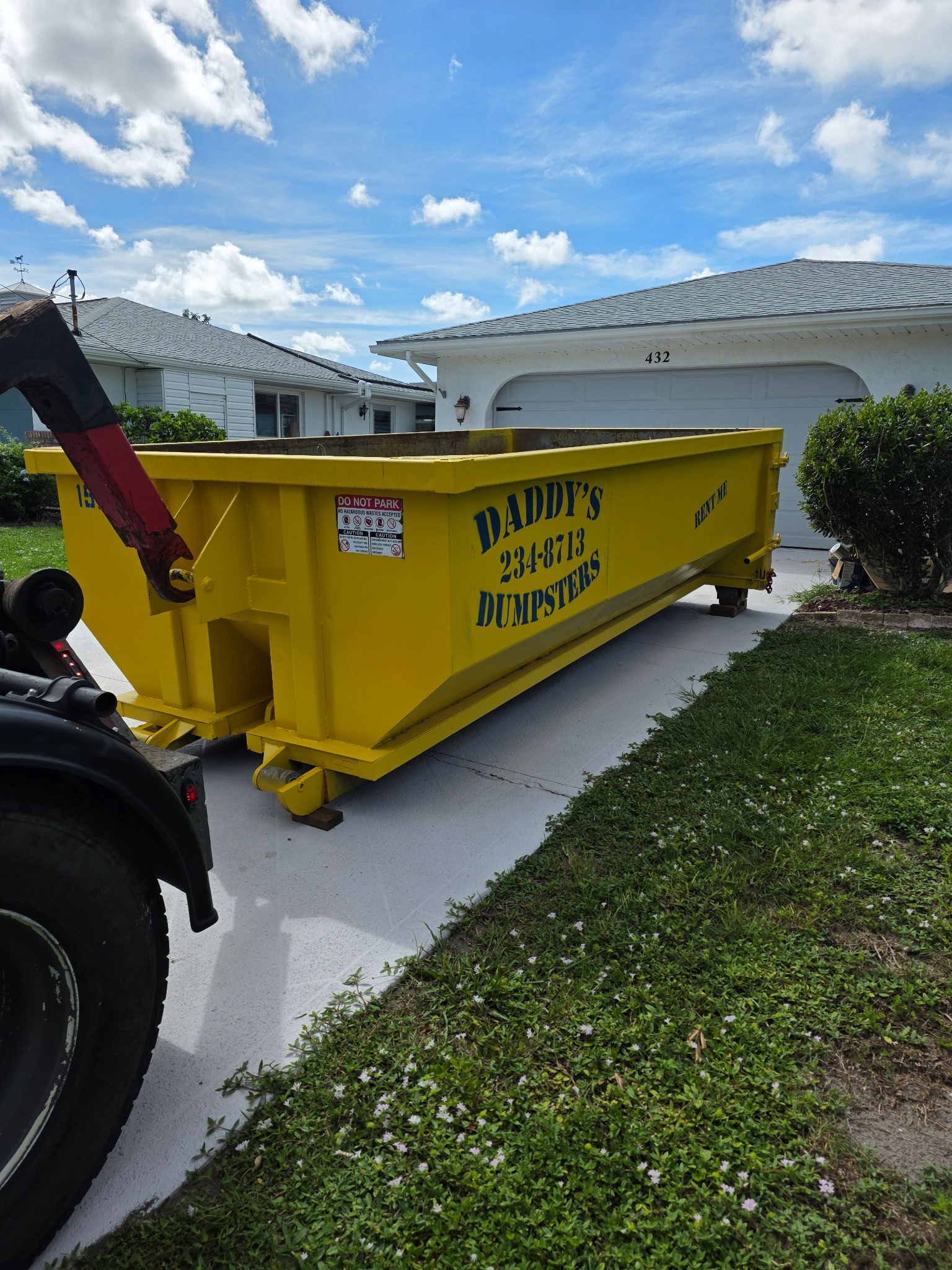 Yellow dumpster on a driveway beside a white house, with a truck and blue sky in the background