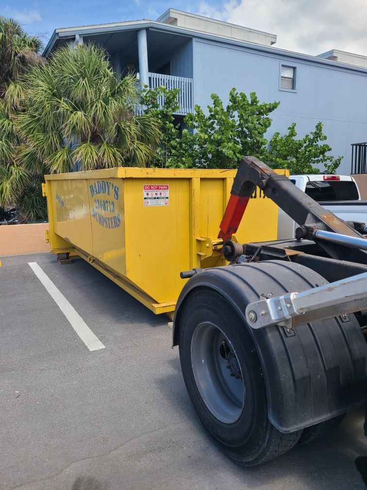20-yard yellow dumpster being moved by a truck in a parking lot beside a light-blue building