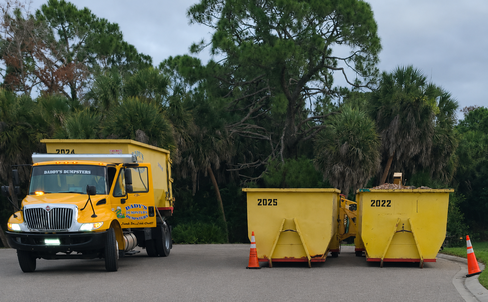 Yellow municipal truck beside two yellow dumpsters and orange cones on a tree-lined road