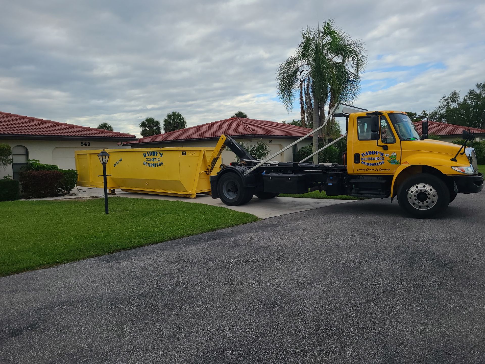 Yellow dump truck parked in a driveway beside a suburban house and lawn