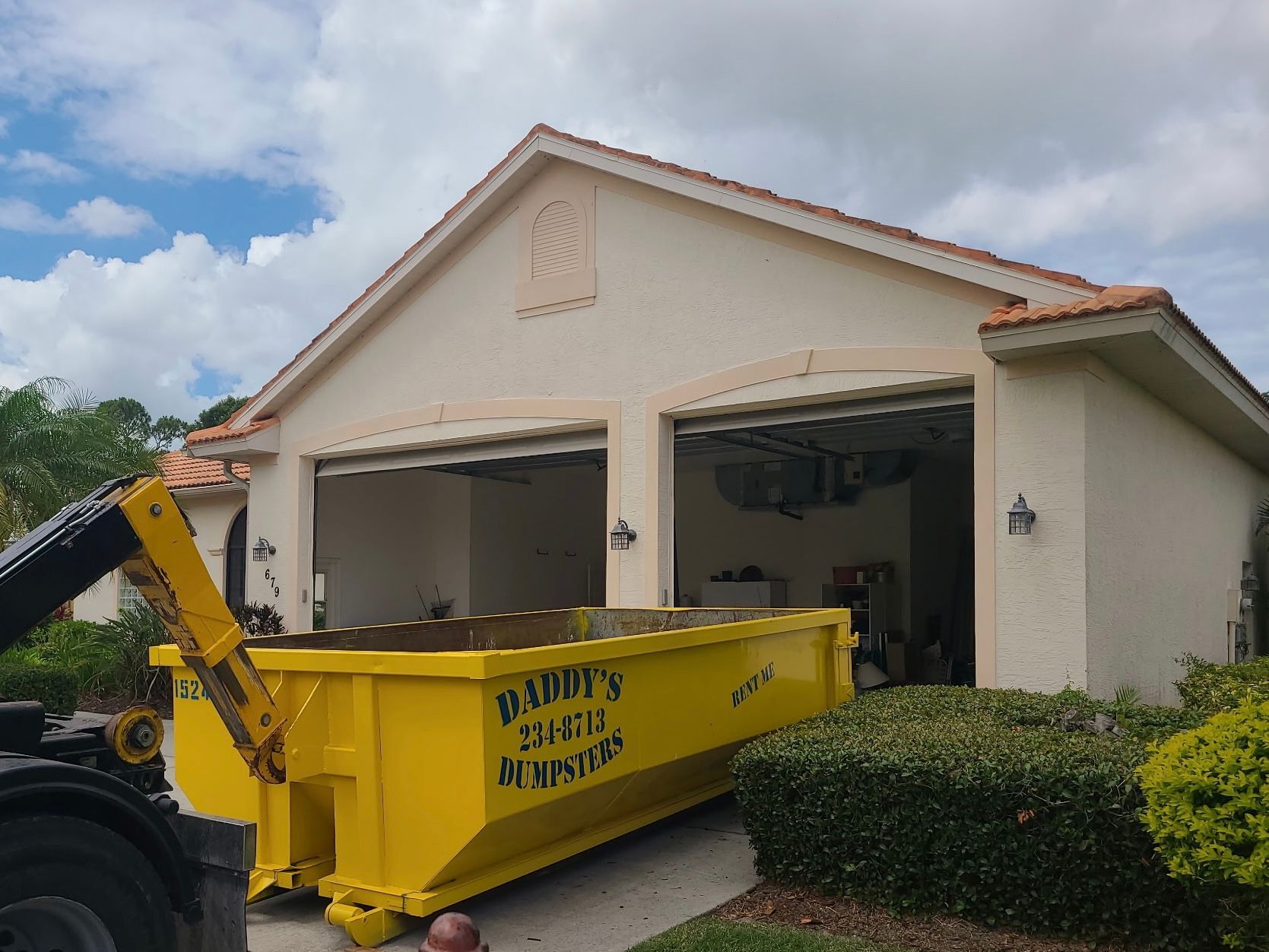 Yellow dumpster in front of open garage of a beige house under a cloudy sky