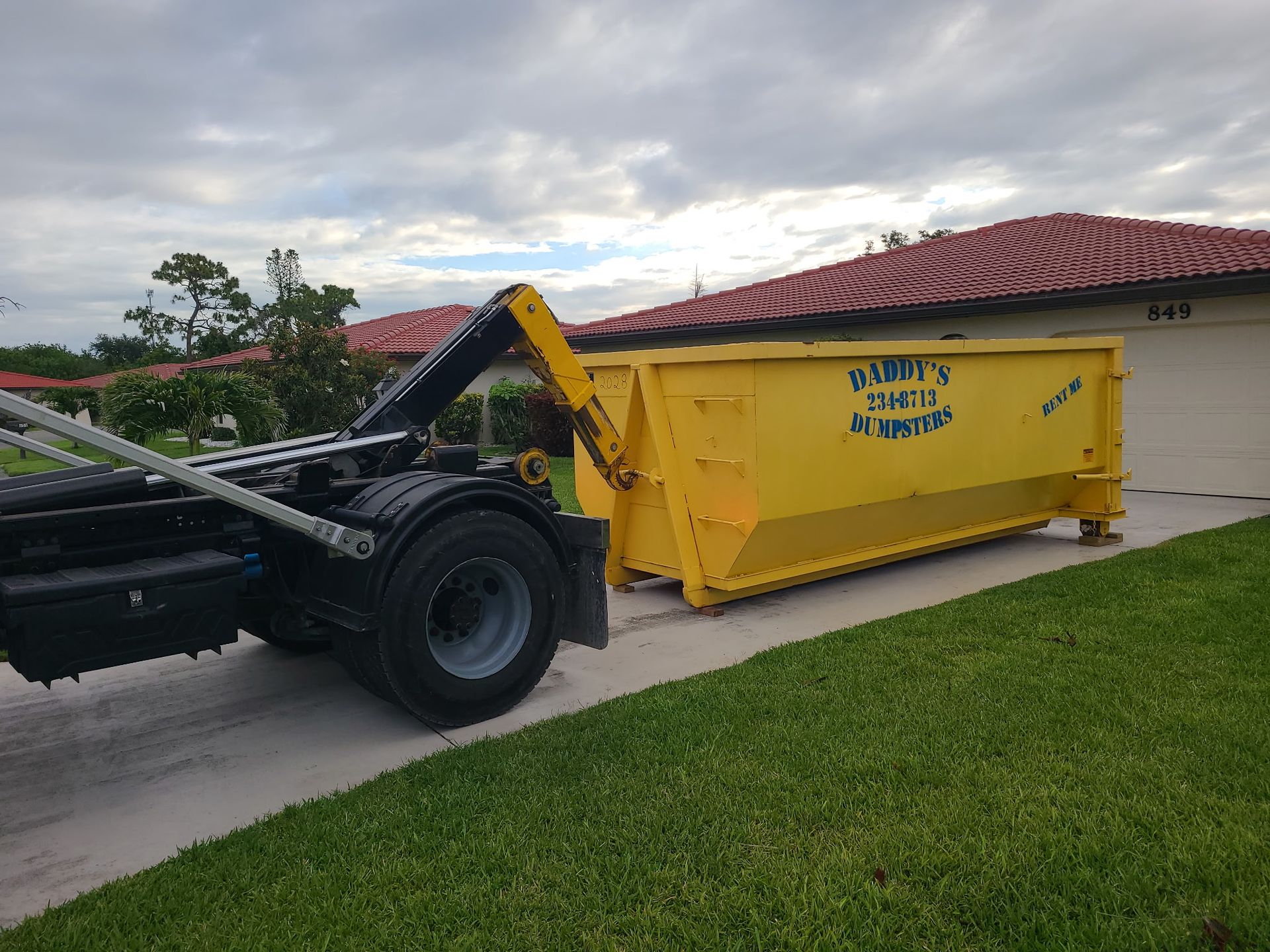 Black tractor arm loading a yellow dumpster beside a suburban house with a red tile roof.