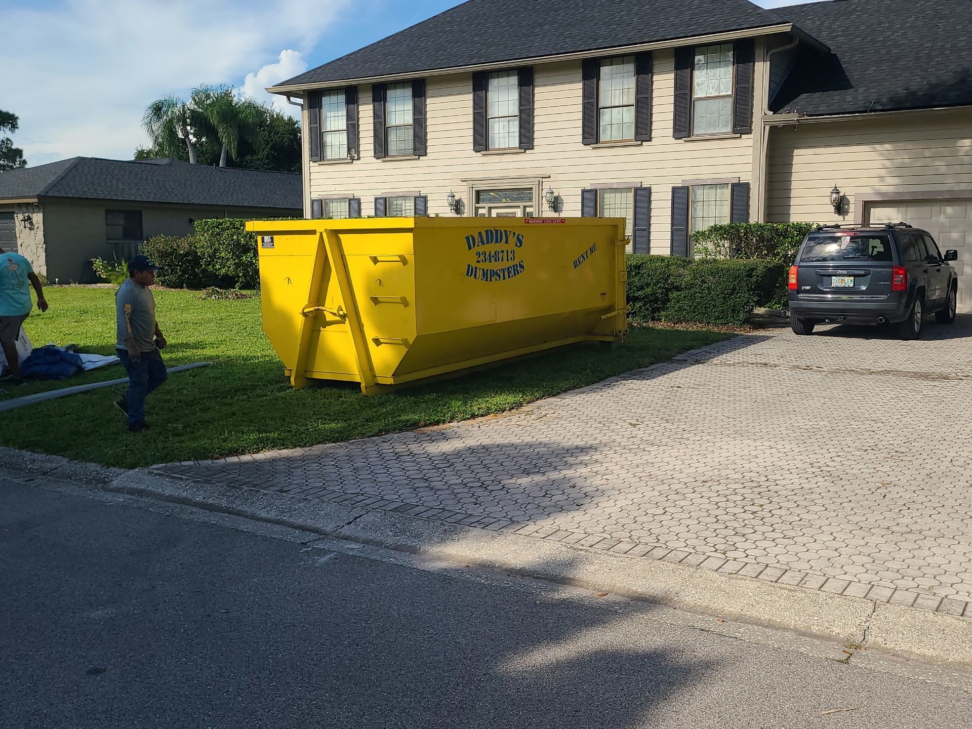 Yellow dumpster on a residential street beside a house, with a person walking nearby and a parked car.