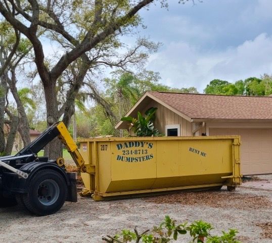 Yellow dumpster beside a house as a front loader empties debris into it under cloudy skies
