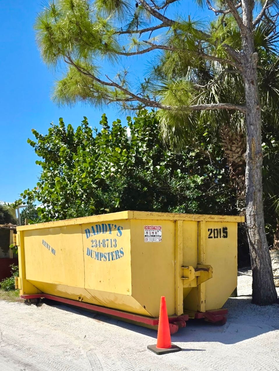 Yellow construction dumpster with orange cone beside it under trees on a sunny day