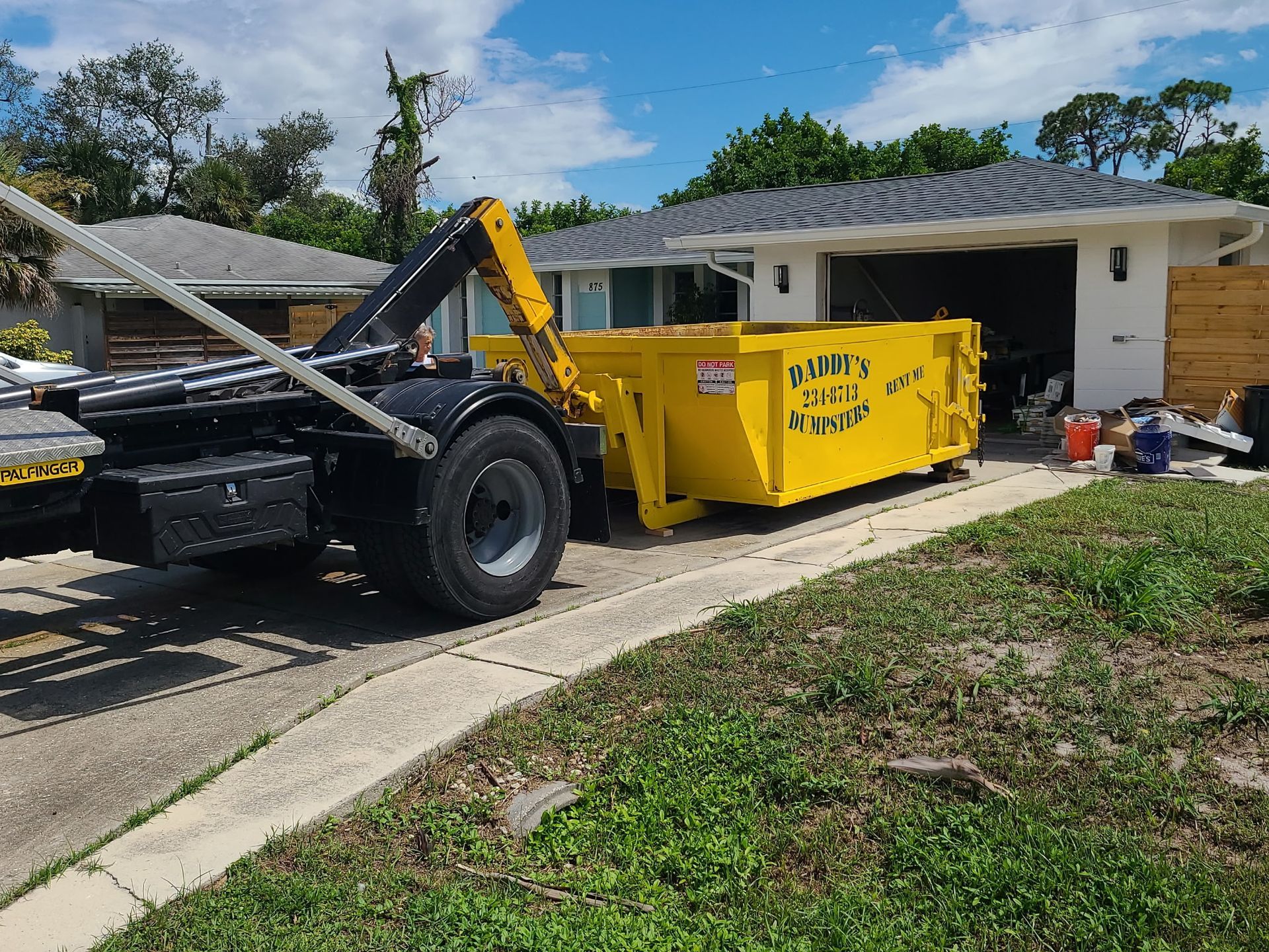 Yellow dumpster on a trailer in a driveway beside a house, with a front loader nearby.
