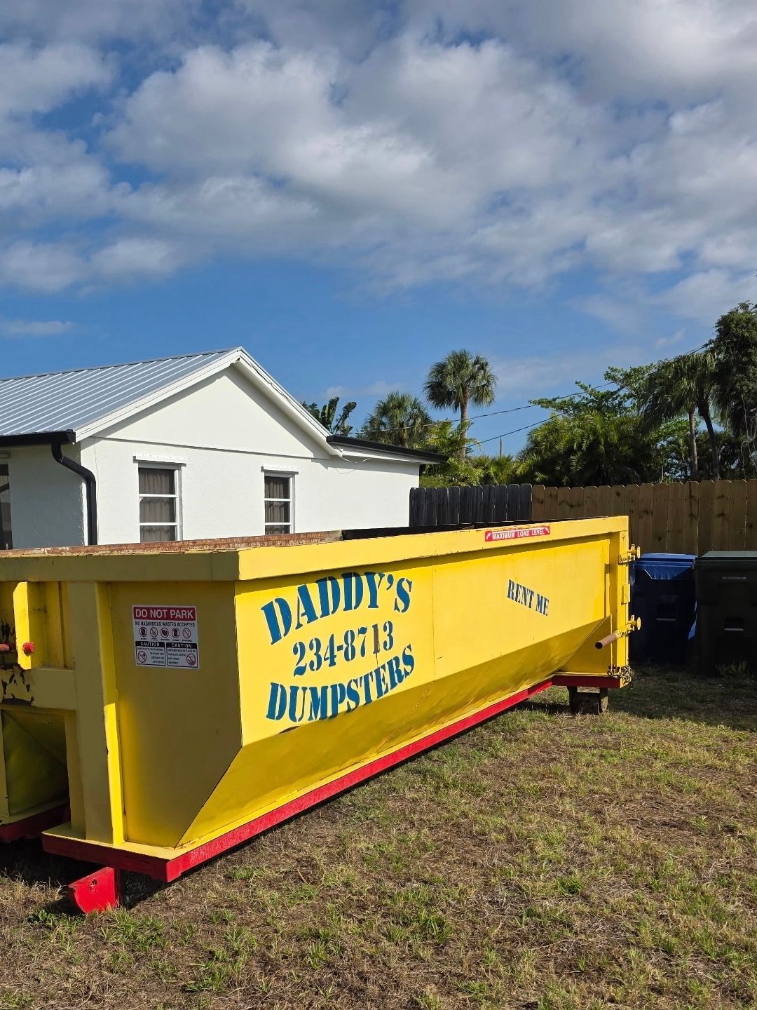 Yellow construction dumpster labeled “Daddy’s Earth Demolition” beside a white house on a sunny day