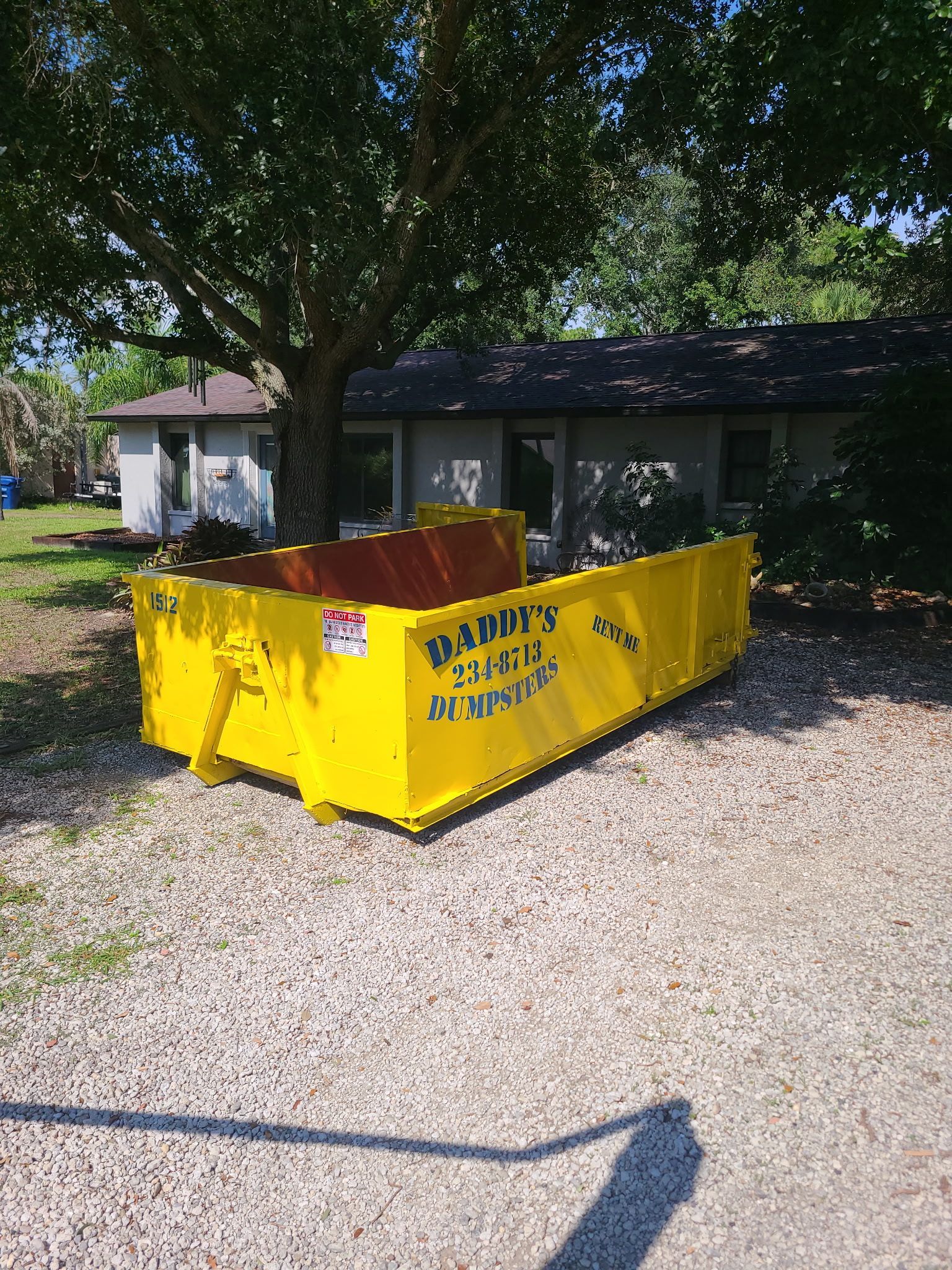 Yellow dumpster in a gravel yard beside a house under a large tree.