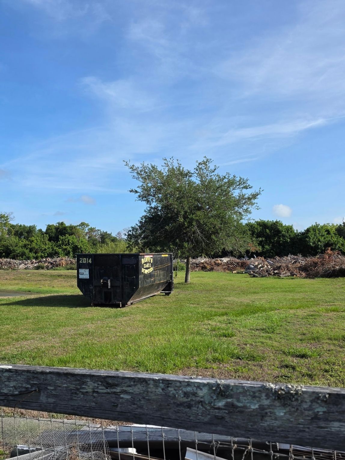 15-yard black dumpster sits on a grassy field under a blue sky, with a tree and rocks in the background.