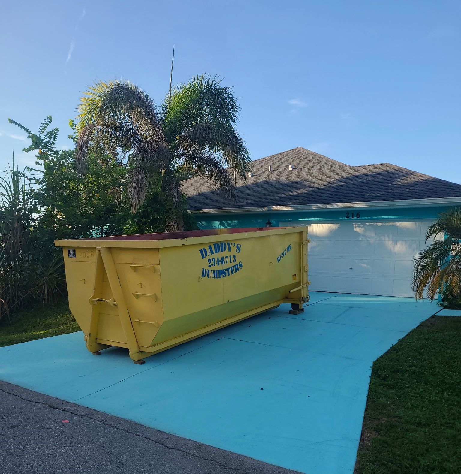 Yellow dumpster on a bright blue driveway beside a house with palm trees and clear sky.