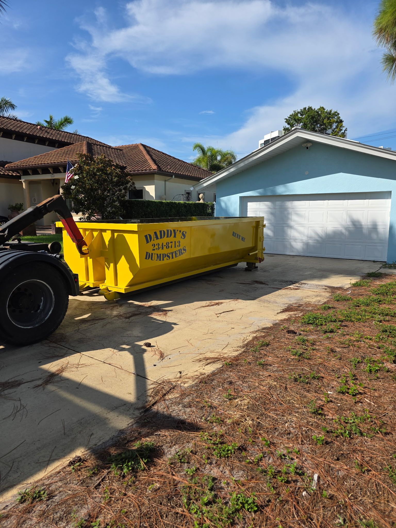 15-yard  yellow dumpster in a driveway beside a light blue garage on a sunny suburban street.