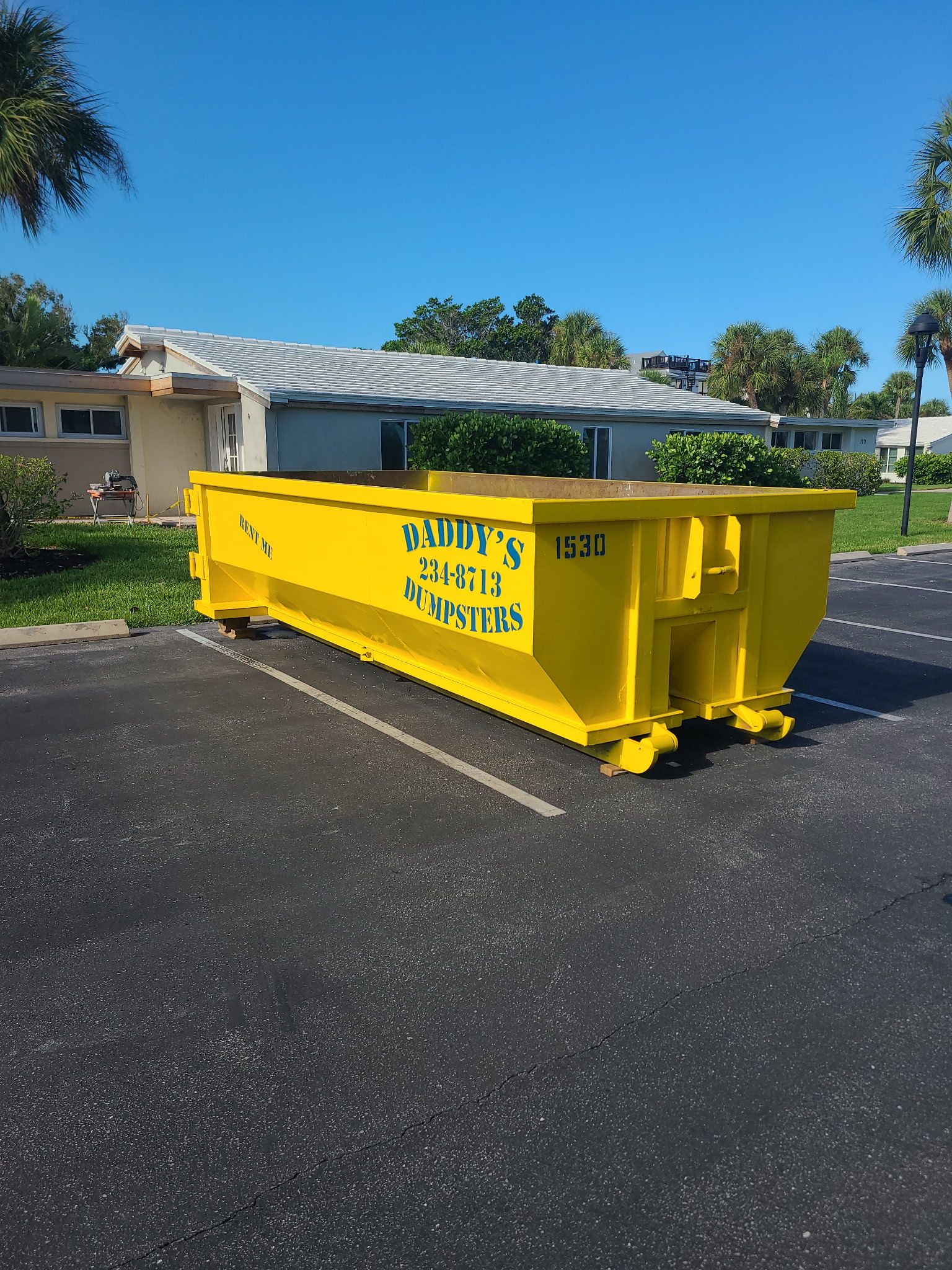 Yellow construction dumpster in a parking lot beside a house, under a clear blue sky.