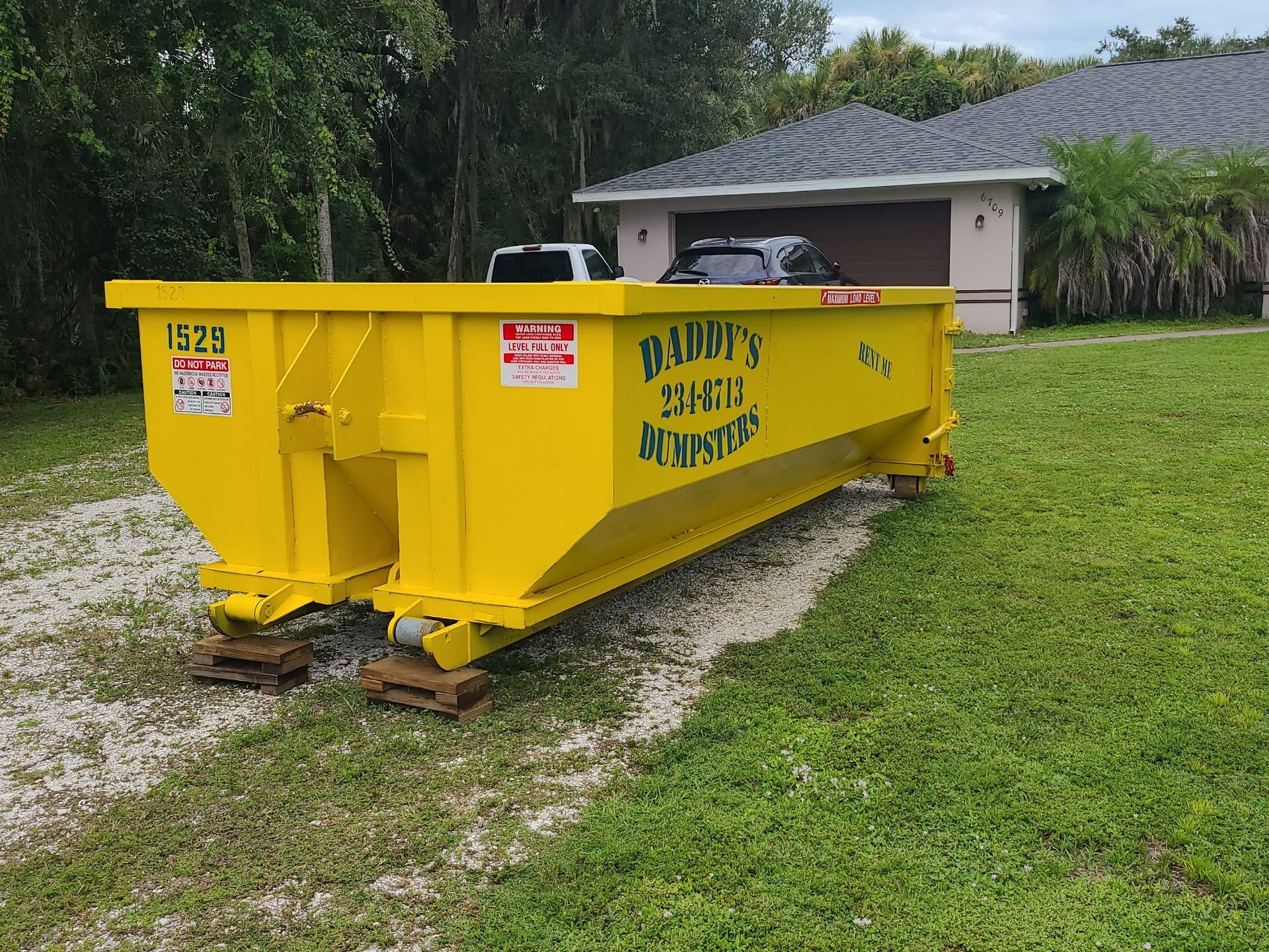 Yellow dumpster on a gravel driveway beside a house and green lawn.