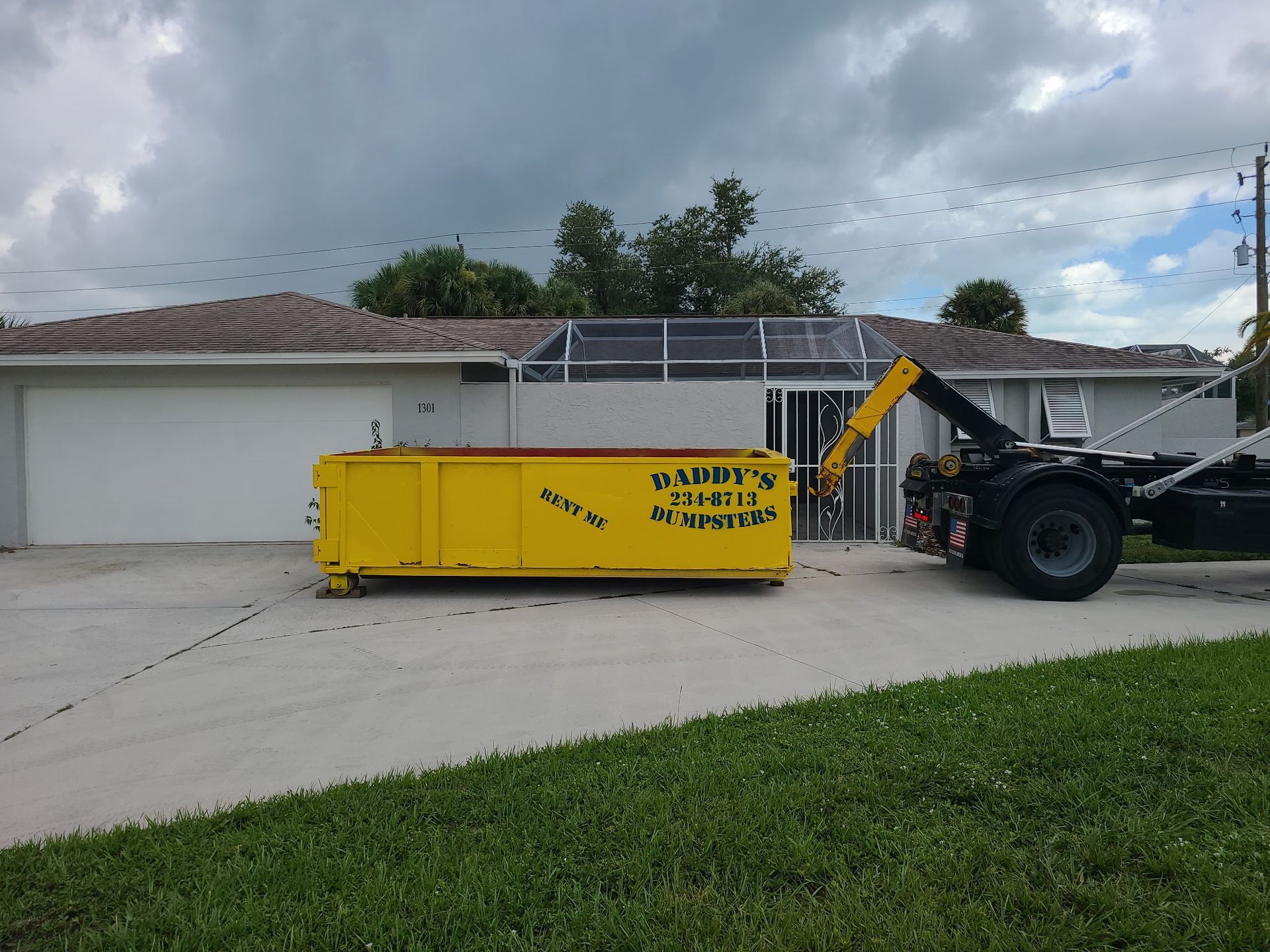 Yellow dumpster truck parked in a driveway beside a house under cloudy skies.