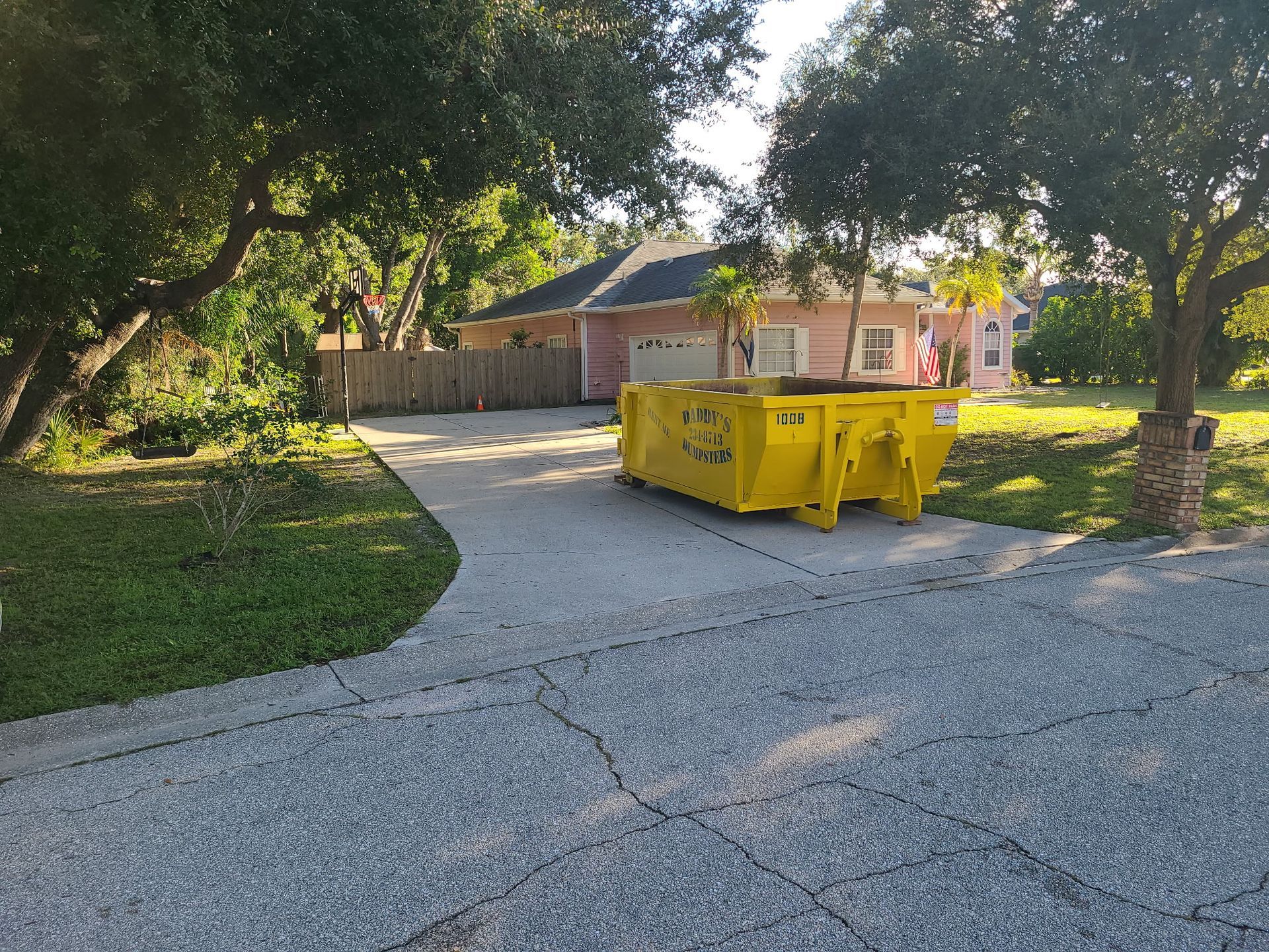 Yellow dumpster on a gravel driveway beside a house, with trees and lawn in the yard.