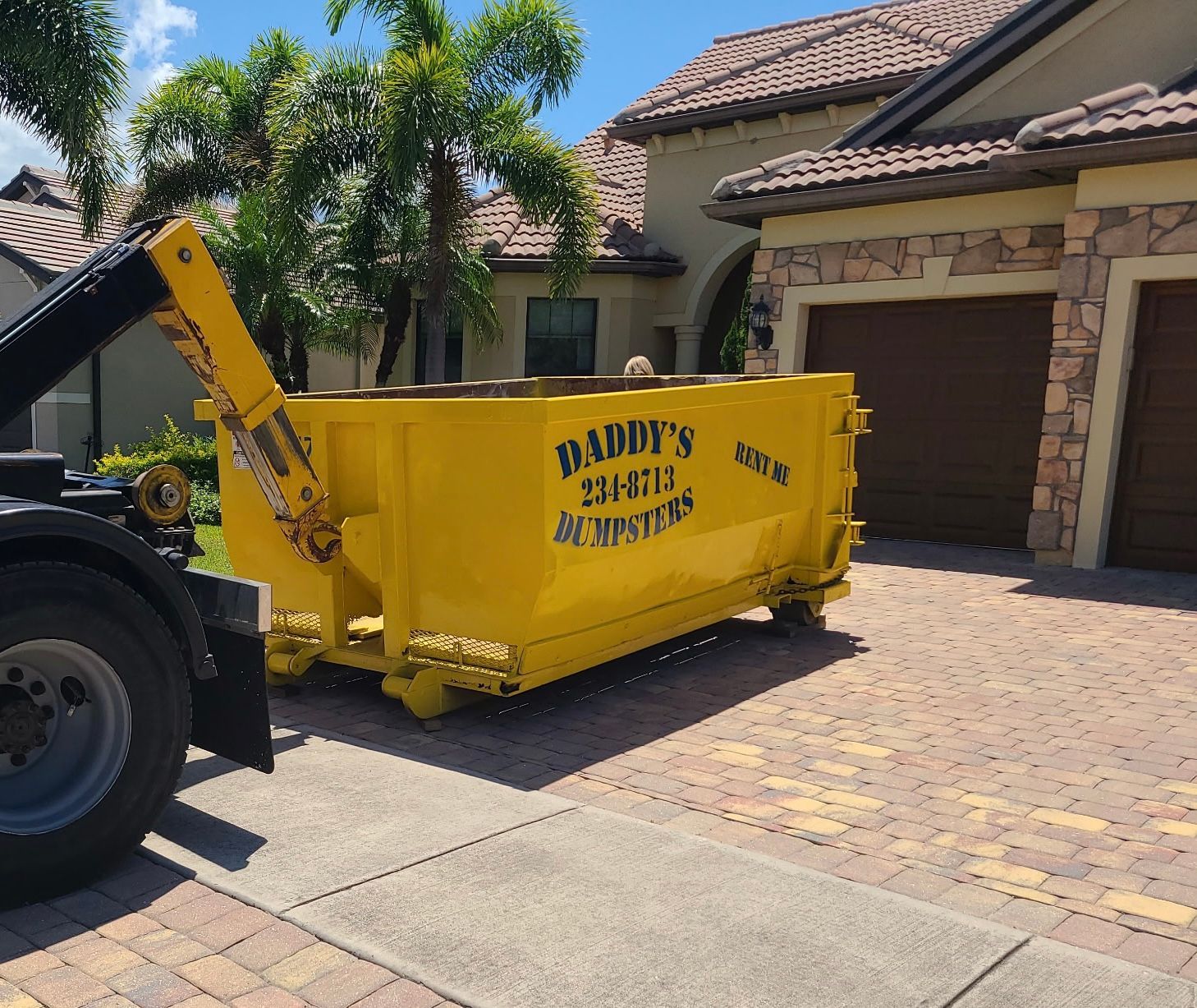Yellow dumpster in a suburban driveway outside a house with a garage and palm trees.