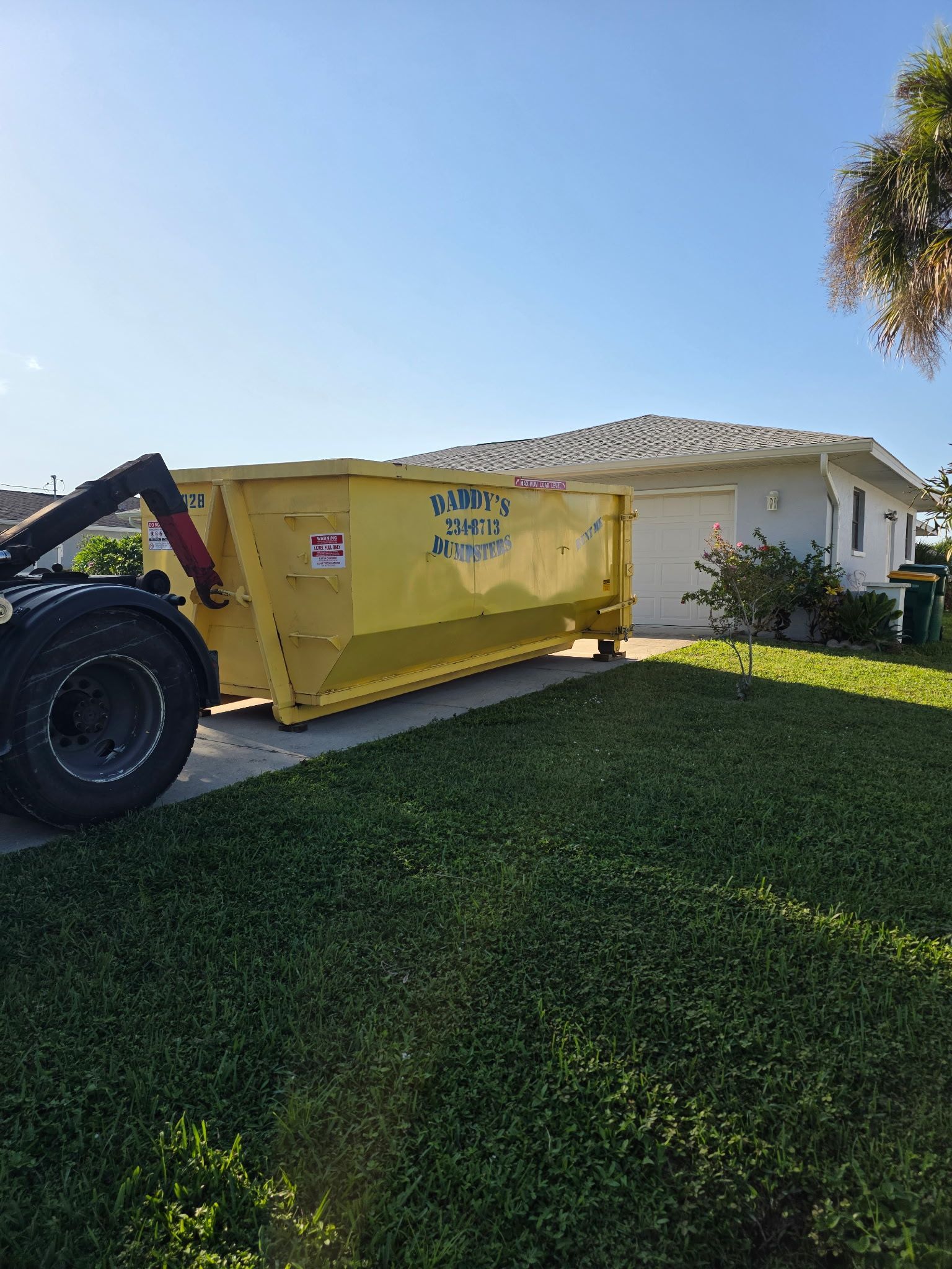 Yellow dumpster on a lawn beside a white house, with a tractor attached under a bright blue sky.