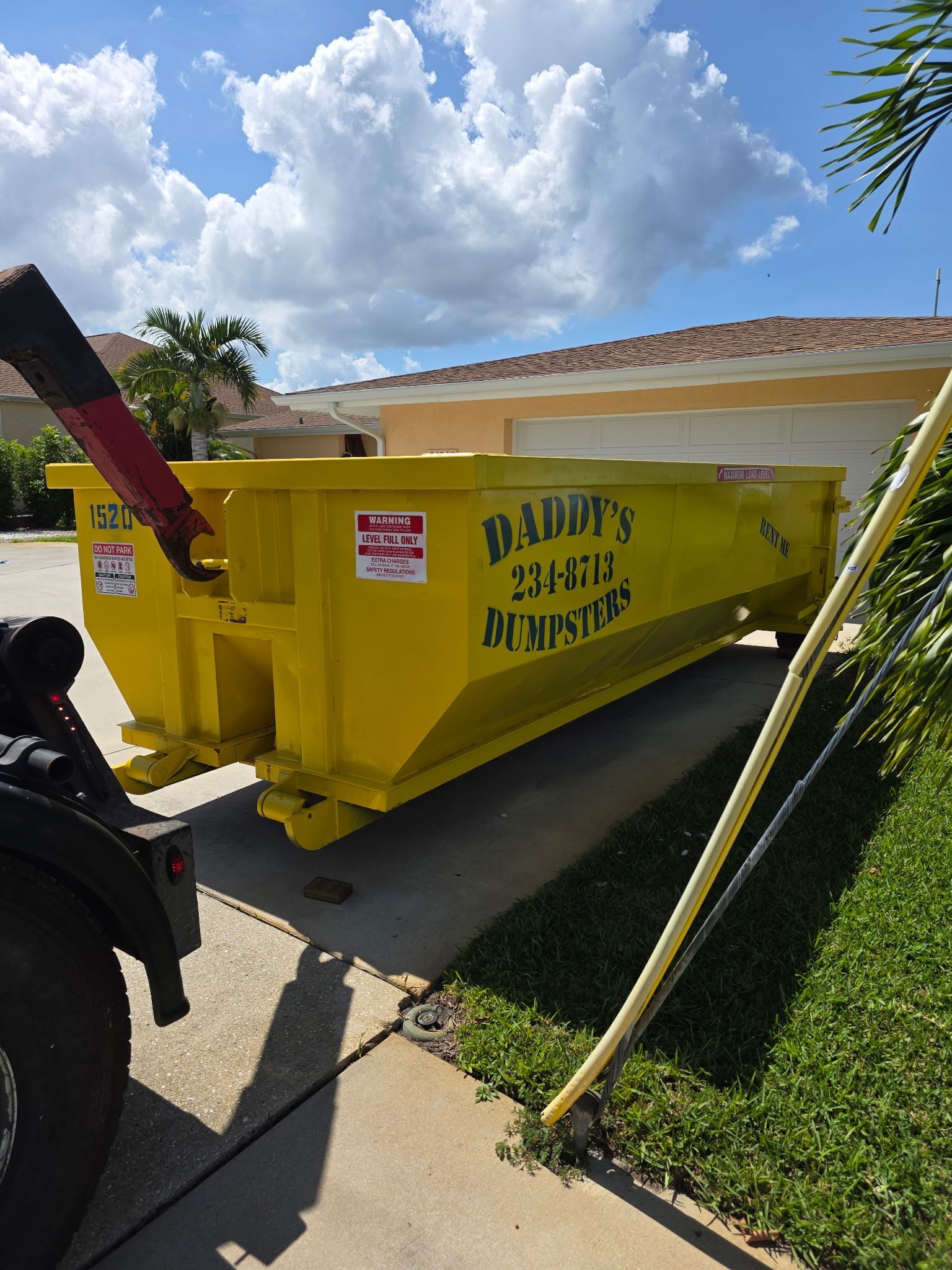 Yellow dumpster on a sunny street beside a truck, with palm trees and a house in the background.