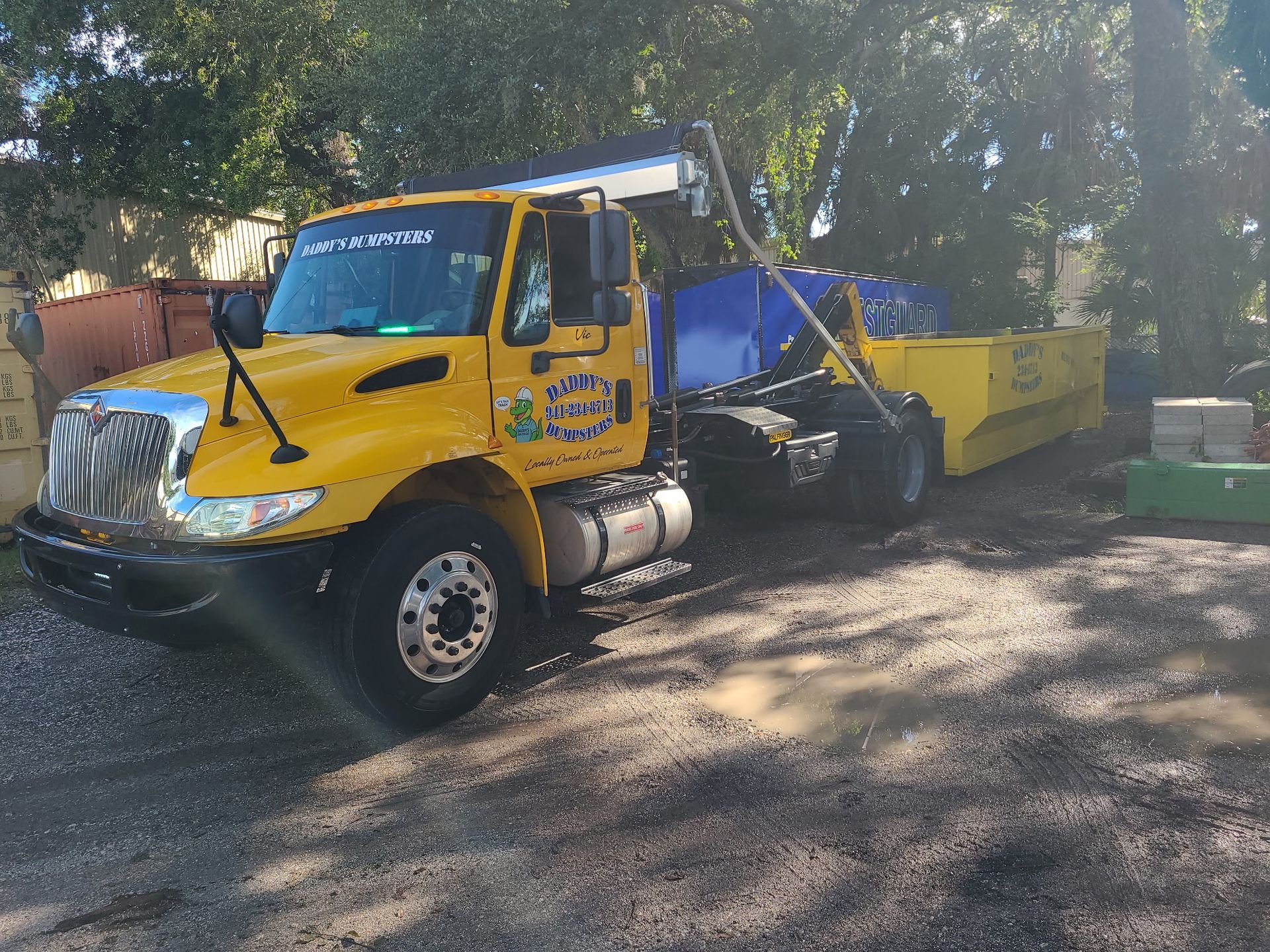 Yellow road paver truck parked on a tree-lined street, with a worker in the cab