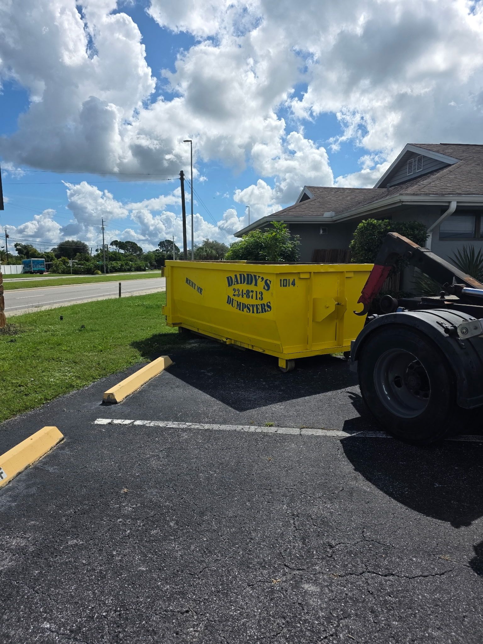 Yellow dumpster on a blacktop parking lot beside a pickup truck and house under a cloudy sky