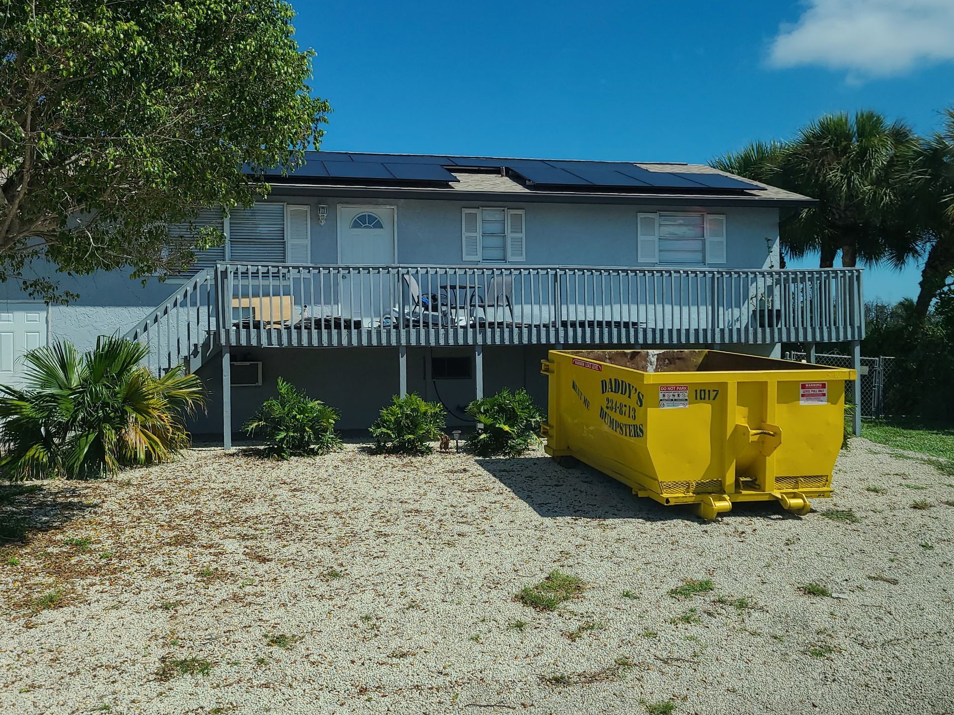 House with white porch and yellow dumpster on gravel driveway under a blue sky