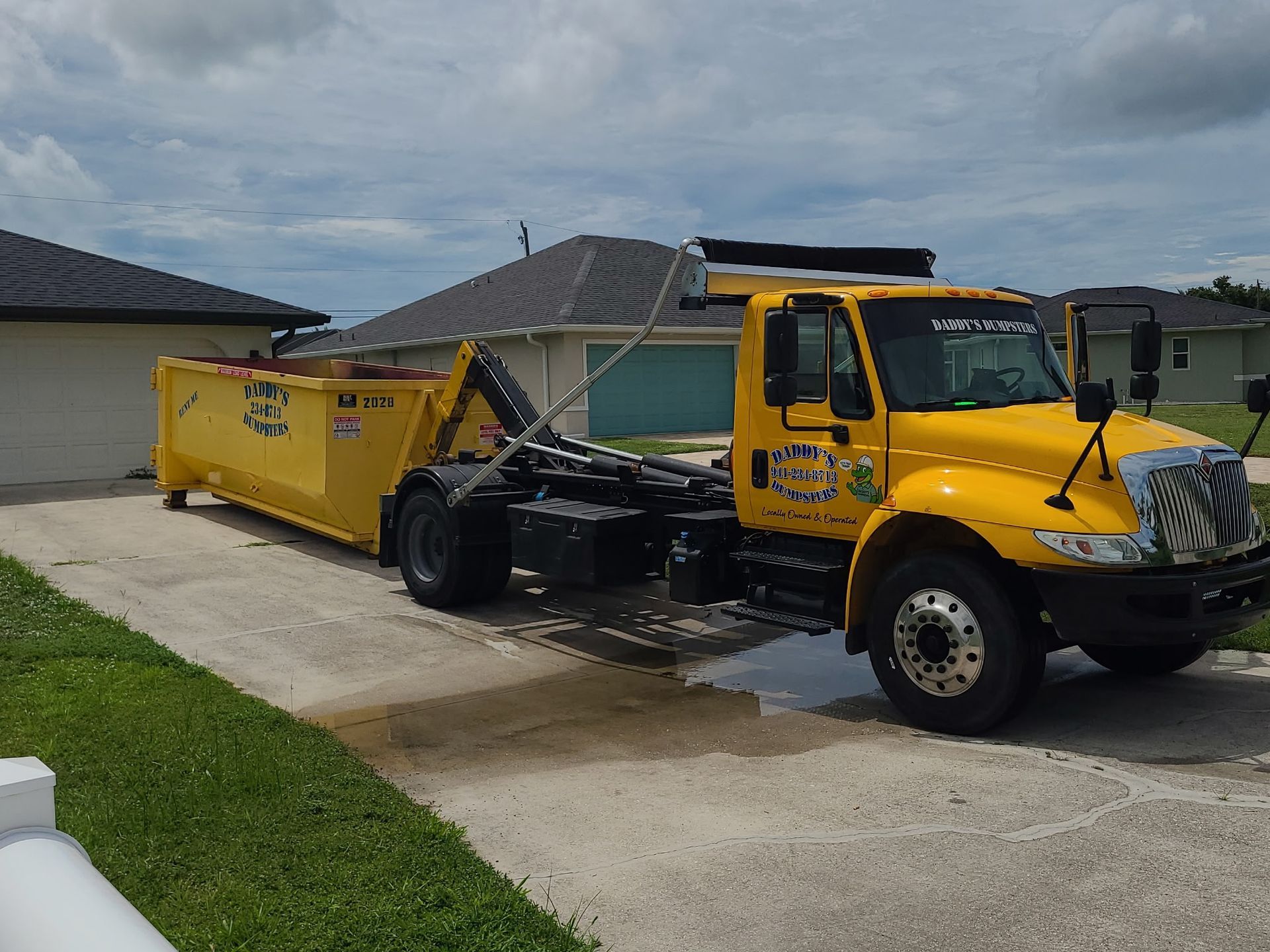 Yellow dumpster truck parked in a driveway beside a house, with a cloudy sky overhead