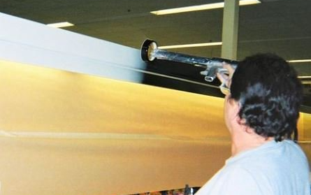 Person spraying a large white sheet of material with a black nozzle in an industrial setting.
