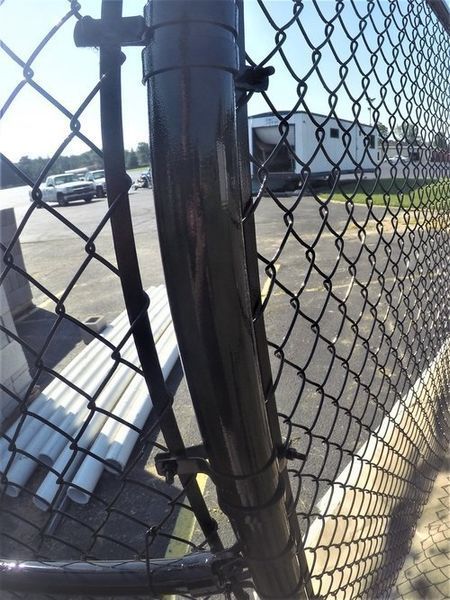 Black chain-link fence with a curved, black metal post secured with black fasteners. Background includes vehicles and a building.