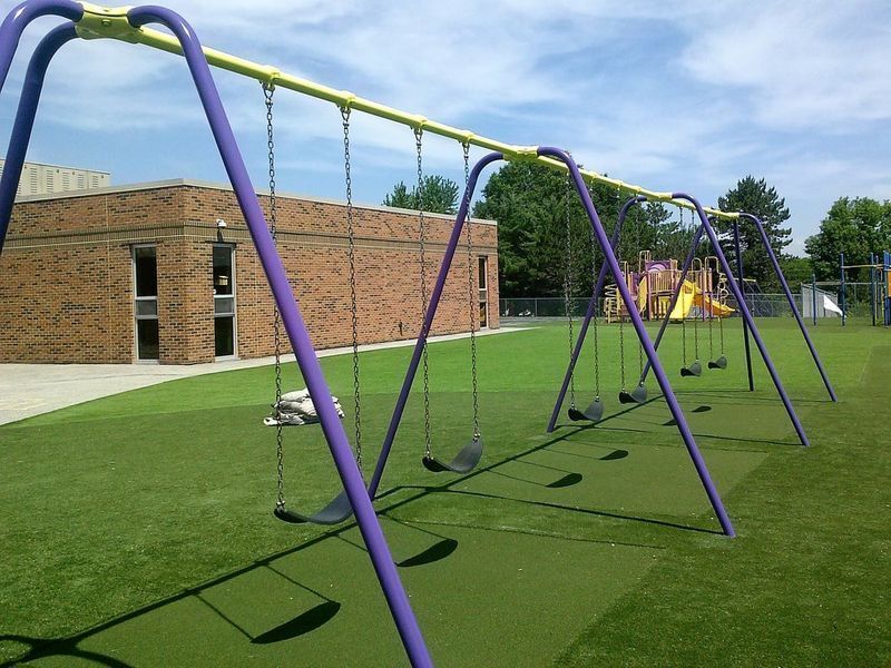 Purple swing set on green ground near a brick building and other playground equipment under a blue sky.