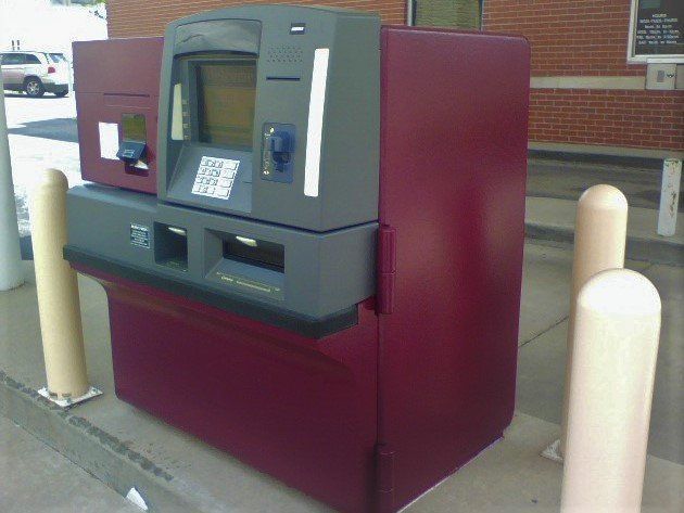 ATM machine, maroon and gray, outdoors, beside sidewalk.