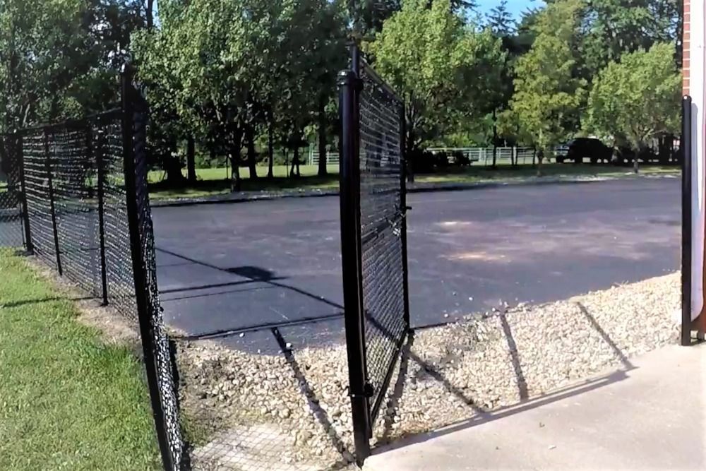Black metal gate open, leading to a paved area with gravel base, surrounded by a fence, and trees in the background.