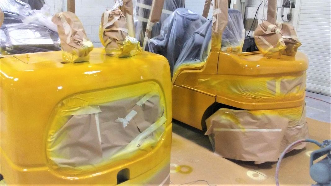 Two yellow forklift cabs being painted in a workshop, masked windows, paper-covered parts.