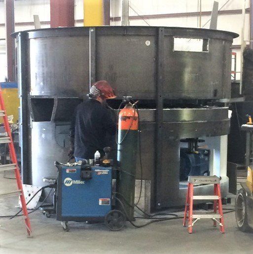 Welder working on large metal industrial machinery with a welding machine and oxygen tank in a warehouse.