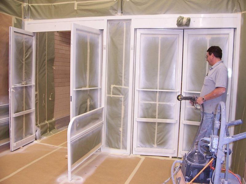 Man spray painting a white door in a room covered with plastic sheeting.