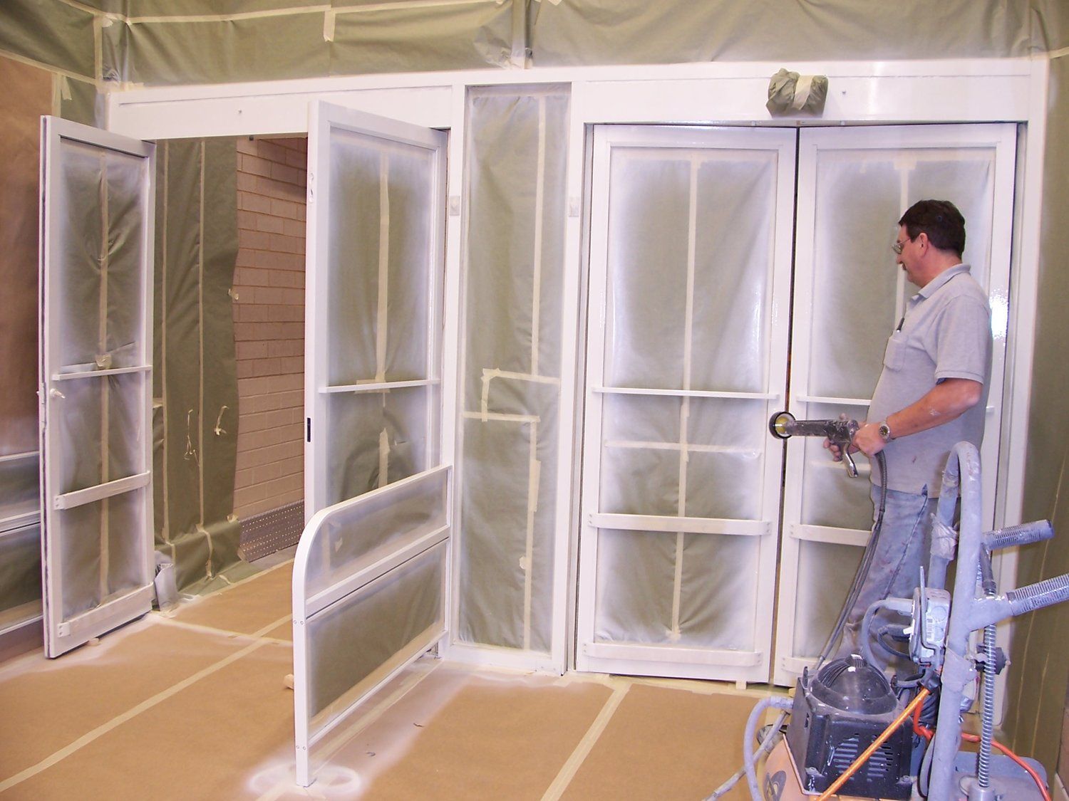 Man painting doors in a spray booth with protective plastic and tape, using a paint sprayer.