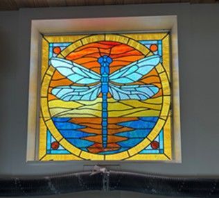 stained glass window: a blue dragonfly within a circular design; orange and yellow sky above blue water