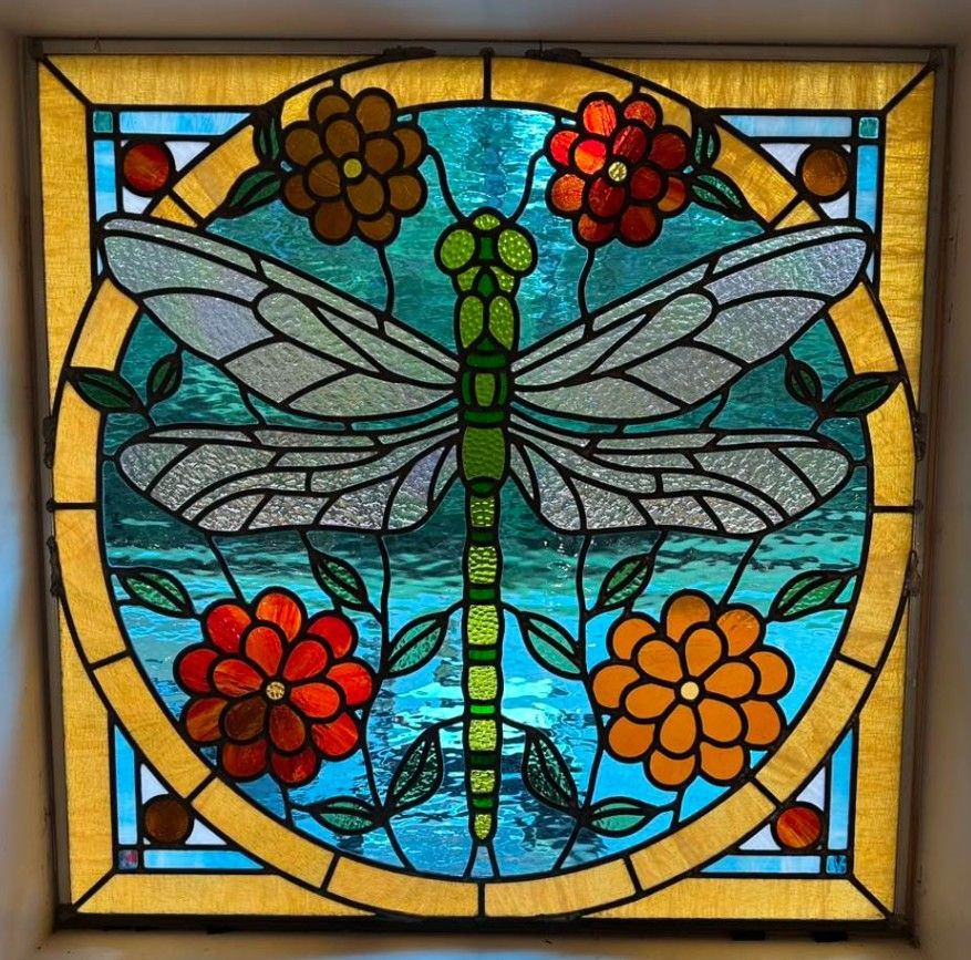 stained glass square with green dragonfly, blue water, and orange and brown flowers on yellow frame