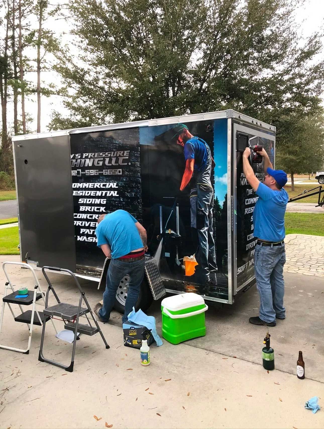 Two men installing graphics on a black trailer, parked outside. One man on a ladder, two drinks sit nearby.
