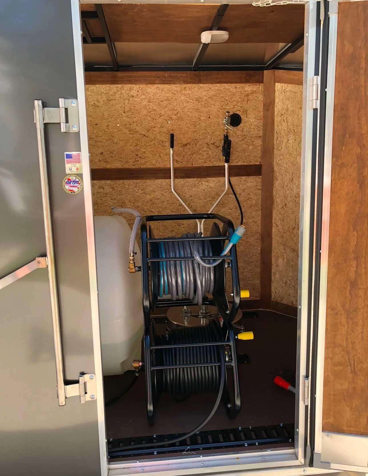 A trailer interior with a hose reel, water tank, and spray nozzle. Door open, wooden walls.