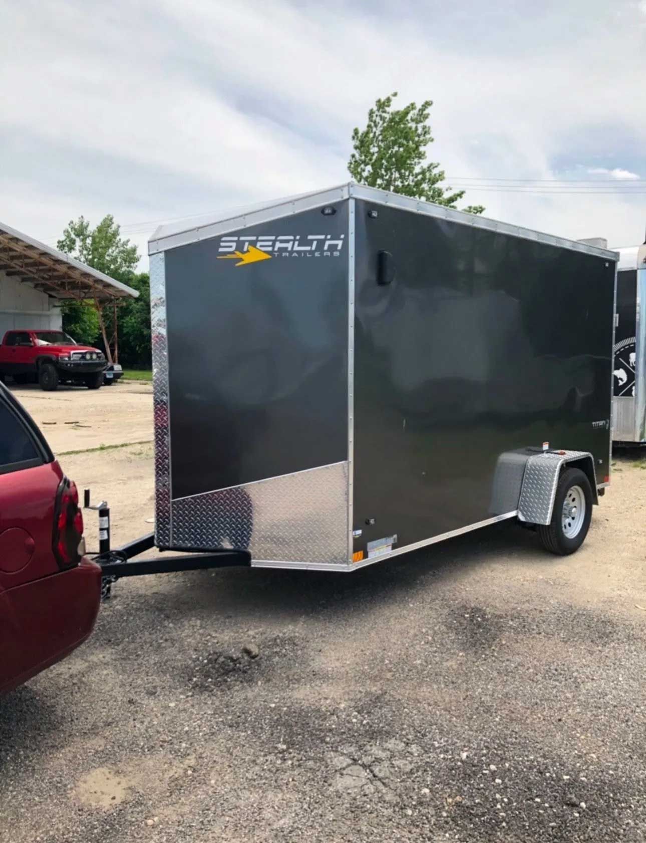 Dark gray Stealth cargo trailer hitched to a red car on a paved lot.