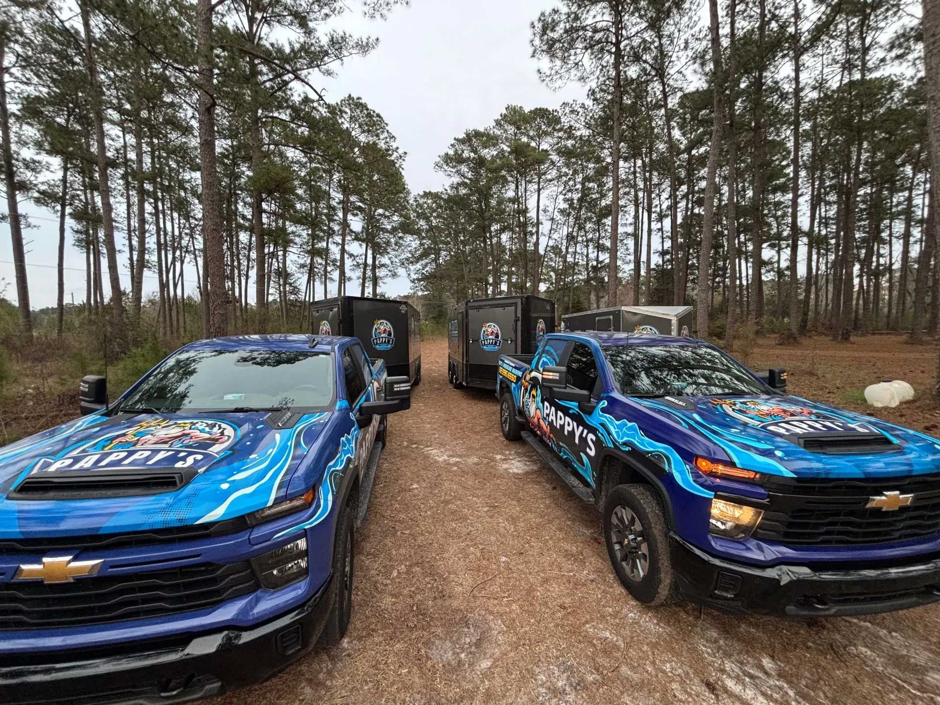 Two blue Chevrolet trucks and a trailer with logos parked on a dirt road in a forest.