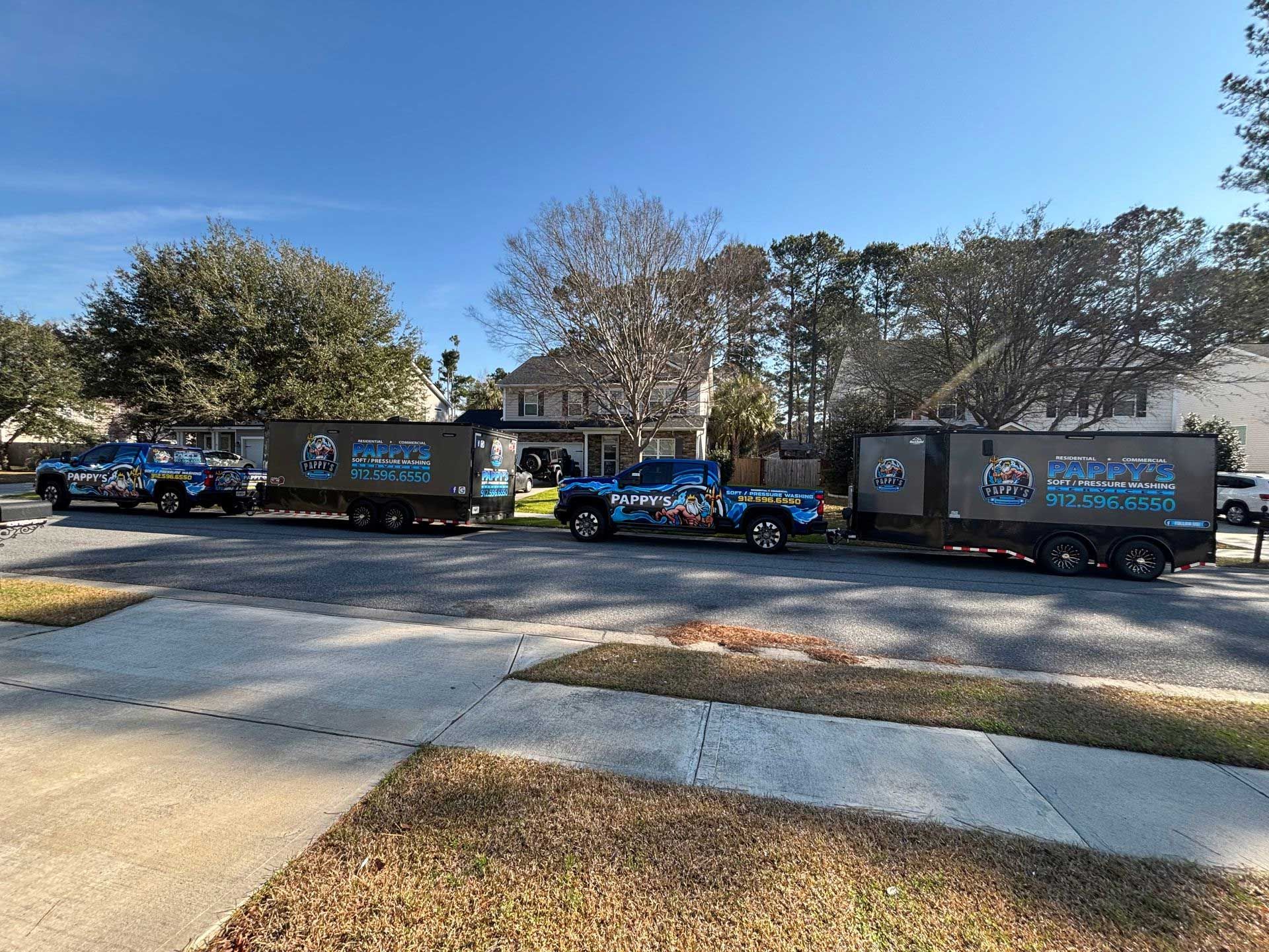 Three blue trucks with trailers parked on a street in front of houses on a sunny day.
