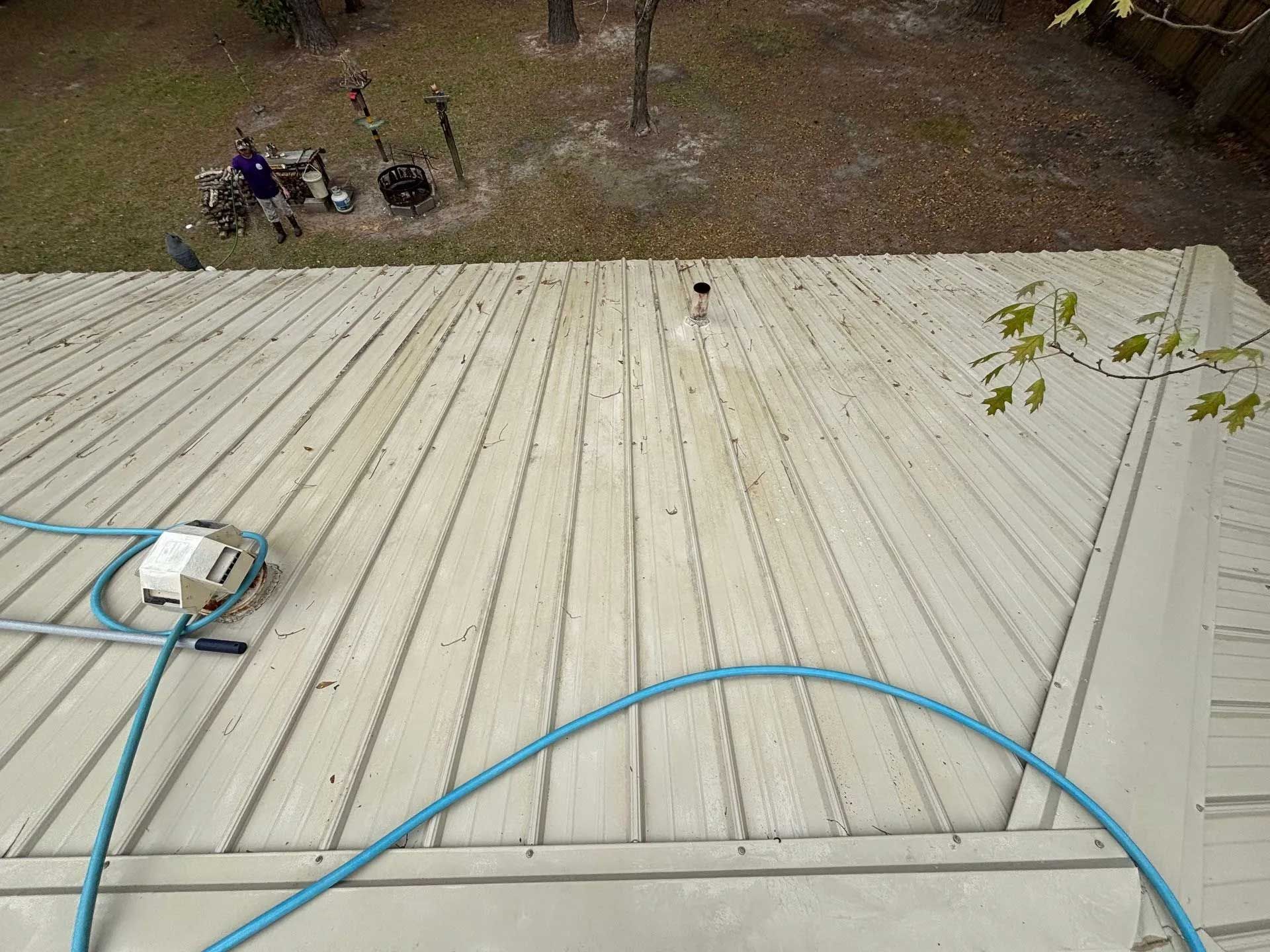 Metal roof being cleaned with a hose. A person is near equipment on the ground in the background.